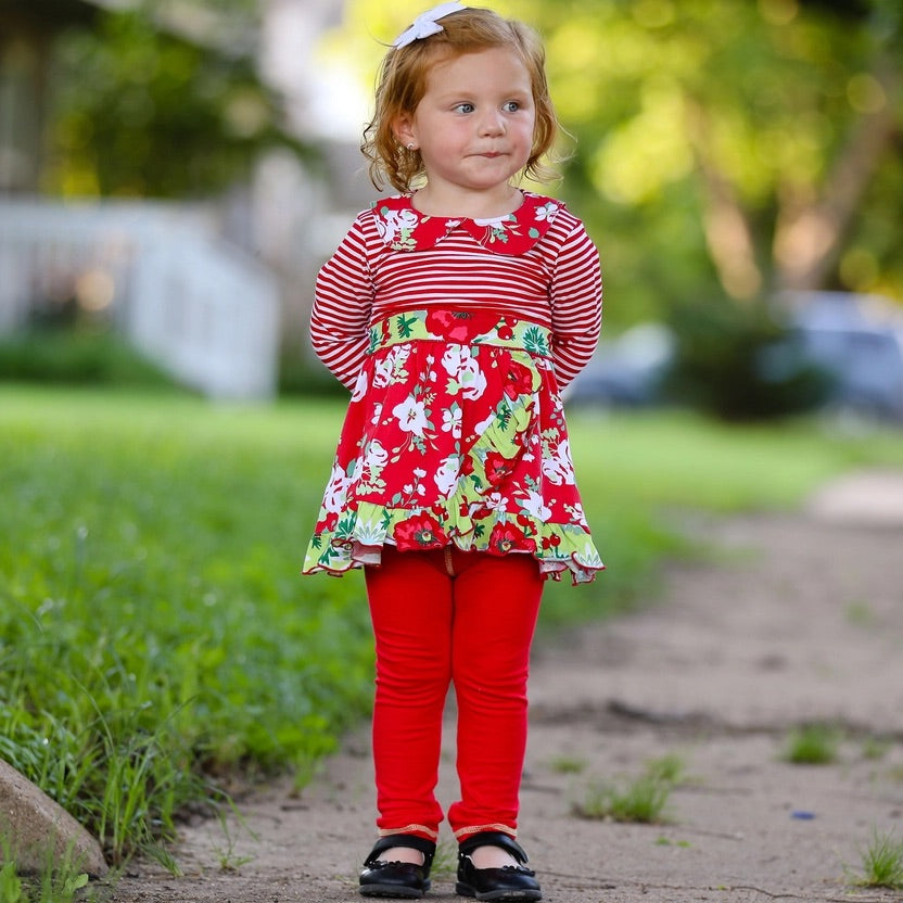 Young girl in a red floral dress and red pants standing outdoors on a grassy path.
