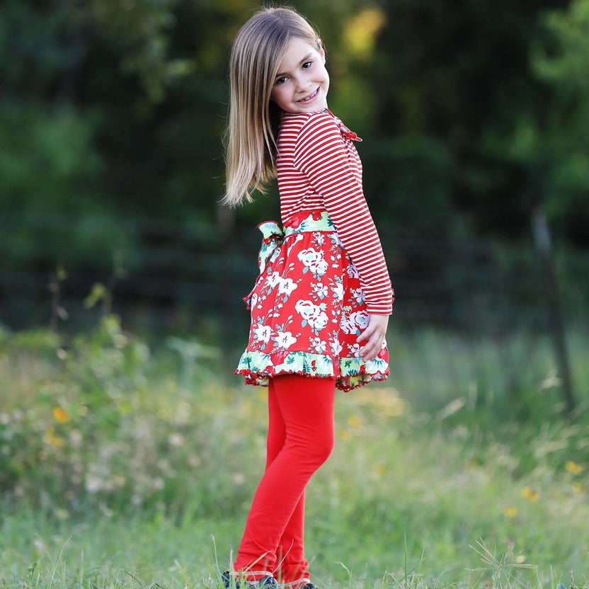 Young girl in a red and white striped dress with floral patterns standing in a grassy field.