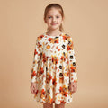 A young girl with light brown hair tied back wears a long-sleeve white dress decorated with a colorful autumn pattern of pumpkins, owls, leaves, and flowers. She is standing against a solid beige background.