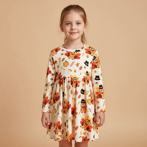 A young girl with light brown hair tied back wears a long-sleeve white dress decorated with a colorful autumn pattern of pumpkins, owls, leaves, and flowers. She is standing against a solid beige background.