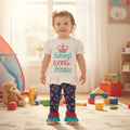 A toddler stands barefoot in a bright children's room wearing a white short-sleeve ruffled top with the words 'Mommy's little Princess' and dark blue patterned leggings with turquoise and pink ruffled cuffs.
