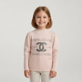 A young girl with shoulder-length brown hair smiling, wearing a light pink ribbed knit sweater with a large Chanel logo and text embellished with silver rhinestones, paired with light beige pants, against a solid light gray background.