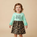 A young toddler girl smiling and wearing a mint green long-sleeve top with 'Wild Child' text and a leopard-print skirt, standing against a solid beige background.