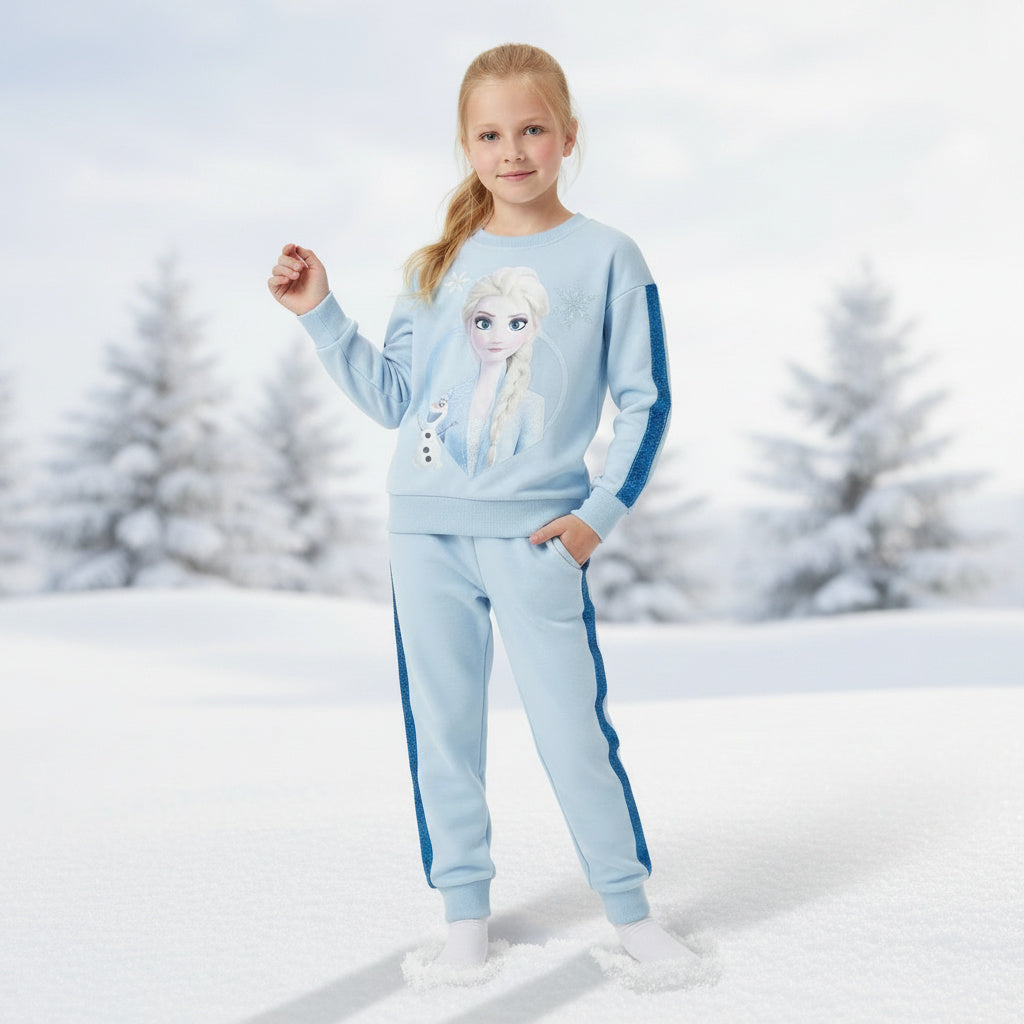 A young girl stands in a snowy outdoor setting wearing a light blue sweatshirt and matching sweatpants with dark blue side stripes and Frozen character graphics.