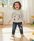 A toddler girl stands barefoot in a bright playroom, wearing a light gray long-sleeve graphic shirt with a cartoon character and the words 'A TINY piece of LOVElessness' paired with dark blue jeans with a pink patch on the knee.