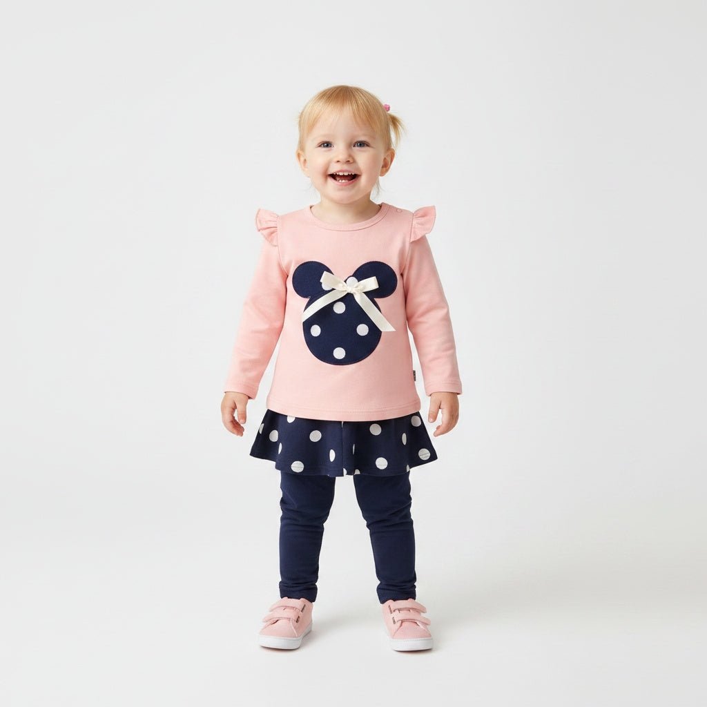A toddler girl stands against a light gray studio background wearing a pink long-sleeve top with ruffled shoulders and a navy polka-dot Minnie Mouse head graphic, paired with a matching navy polka-dot skirt, navy leggings, and pink shoes.