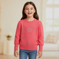 A young girl smiling and wearing a coral pink crewneck sweater with rhinestone-embellished 'CHANEL' logos and blue jeans, set against a softly blurred indoor background.