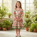 A young girl stands in a bright indoor room with large windows and many potted plants, wearing a long-sleeve tiered dress with a floral print and green trim.
