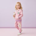 A young girl with blonde hair wearing a pink two-piece tracksuit with bunny patches and a cartoon bunny graphic on the chest, holding a plush bunny toy against a pastel lavender studio background.