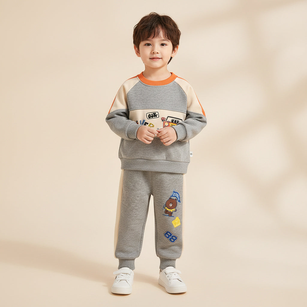 A young boy stands against a warm beige background wearing a gray two-piece sweatshirt and pants set with orange, cream, and cartoon graphics.