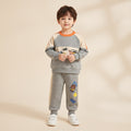 A young boy stands against a warm beige background wearing a gray two-piece sweatshirt and pants set with orange, cream, and cartoon graphics.