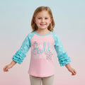A young girl smiling and wearing a 3/4 sleeve raglan t-shirt with a pink body, light blue sleeves with white polka dots, and ruffled cuffs. The front features the text 'Baby, World Outside' in light blue and gray with snowflake graphics.