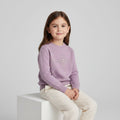 A young girl with long brown hair sits on a white block, wearing a lavender crewneck sweater with a pearl-embellished 'S' logo on the chest and cream-colored pants, against a light gray studio background.