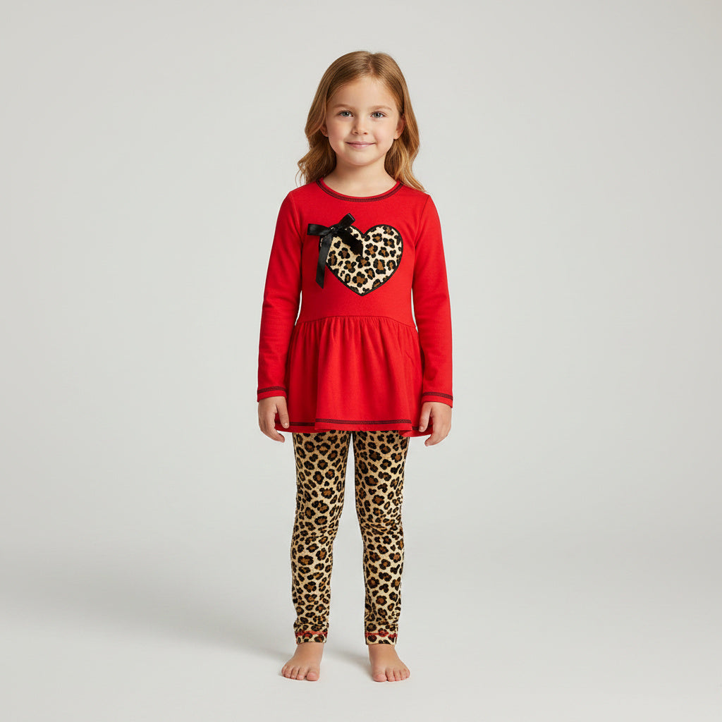 A young girl stands barefoot against a light gray studio background, wearing a bright red long-sleeve top with a leopard-print heart and black bow on the chest, paired with matching leopard-print leggings.