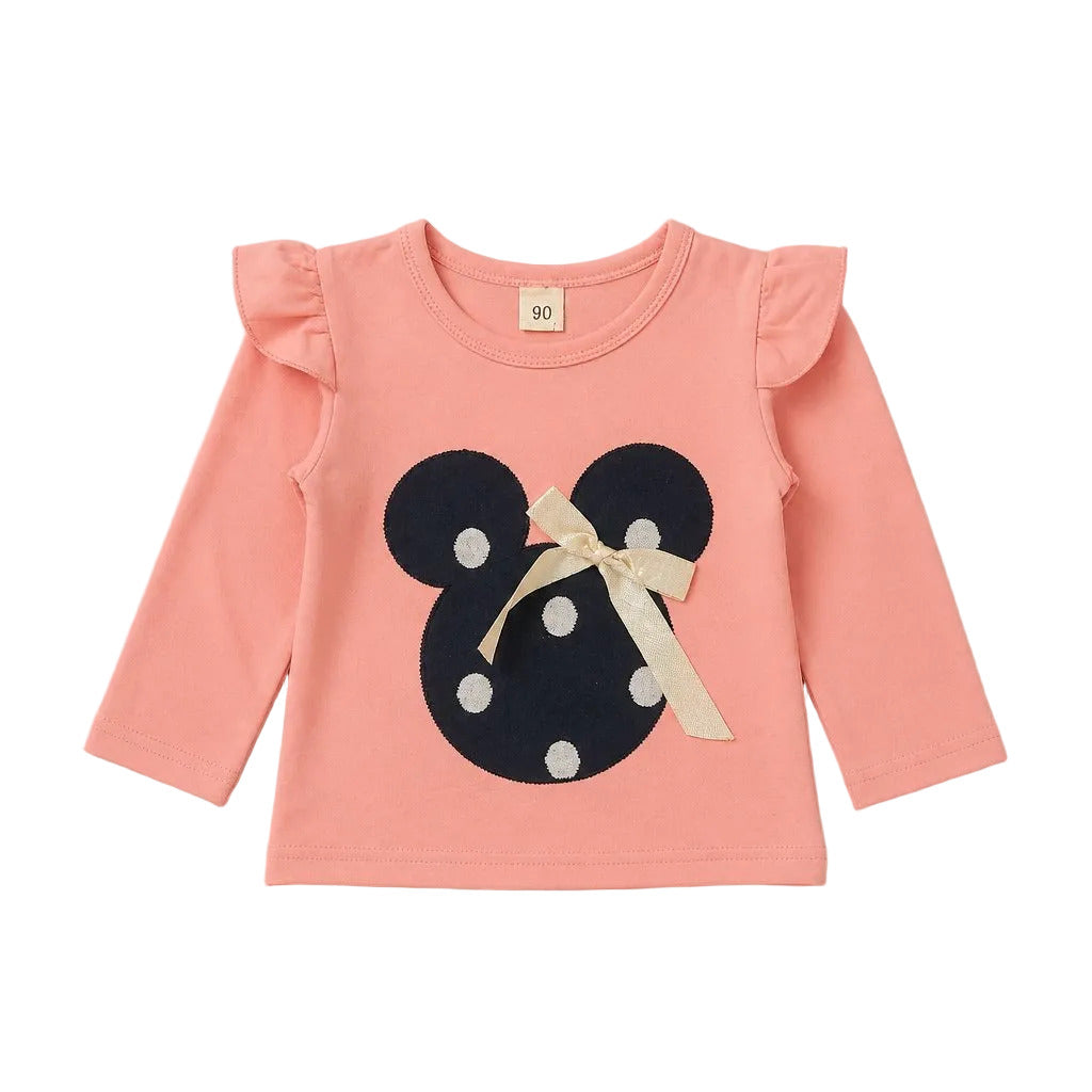 A toddler's long-sleeve top in a soft peach-pink color, featuring ruffled shoulders and a large navy blue Minnie Mouse-style mouse head patch with white polka dots and a cream ribbon bow on the front.
