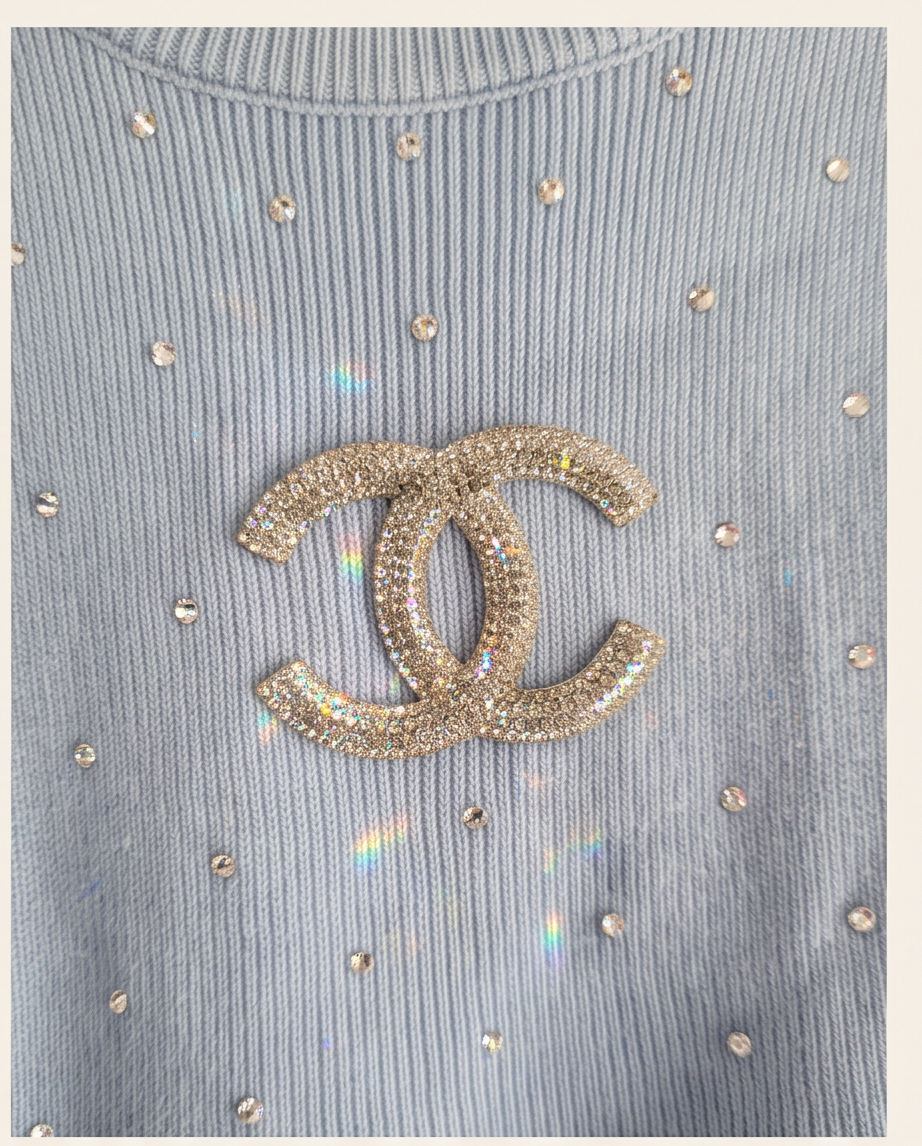 A close-up of a light blue ribbed knit top featuring a large Chanel logo brooch embellished with sparkling crystals, surrounded by scattered rhinestones on the fabric.