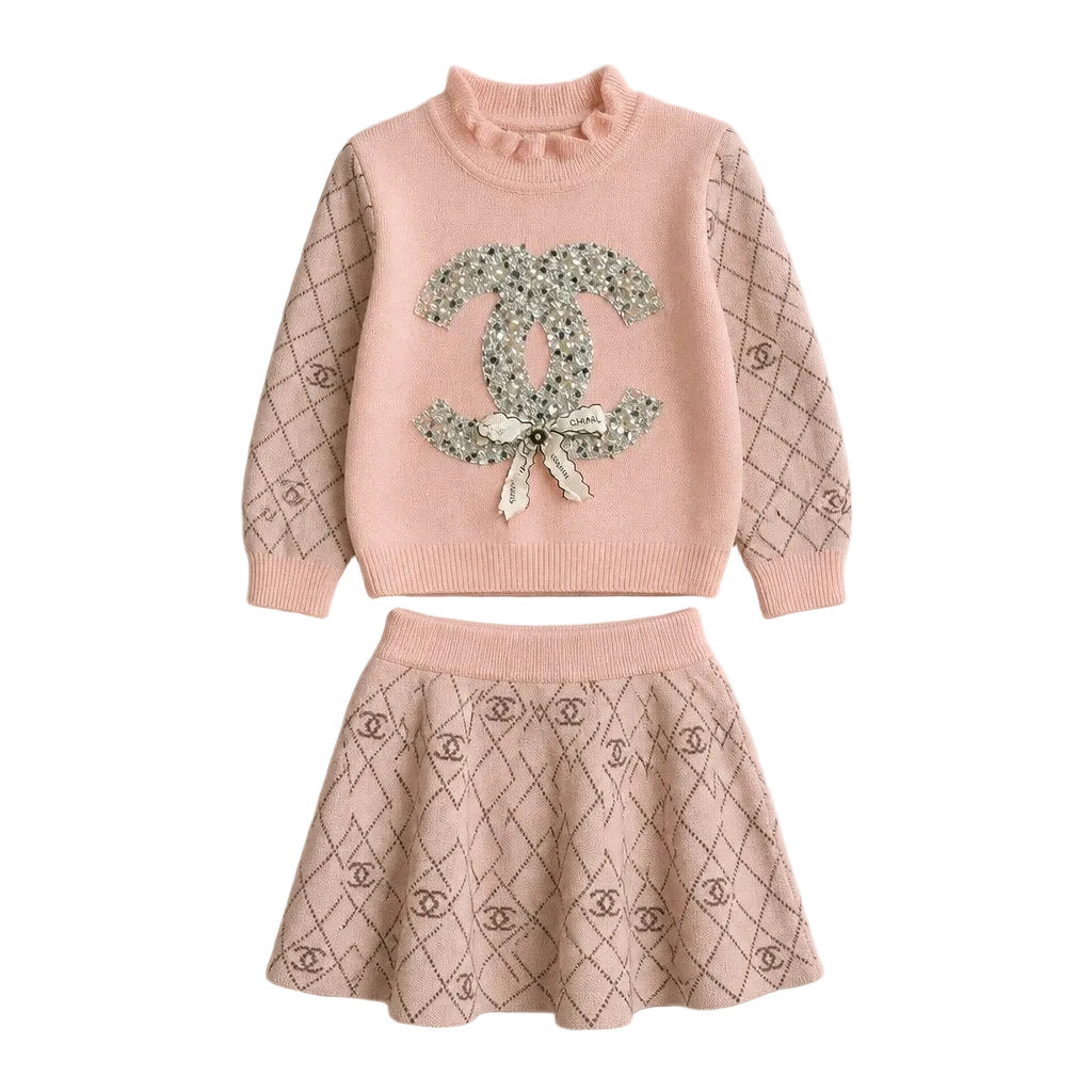 A two-piece girls' knit set in a soft blush pink, featuring a long-sleeve sweater with a floral embellished Chanel logo on the front and a matching patterned skirt.