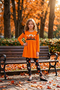 Young girl in an orange dress with a pumpkin design standing on a wooden bench in an autumn park.