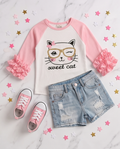 A children's outfit flat lay featuring a white and pink raglan long-sleeve shirt with a cat face graphic and 'sweet cat' text, paired with light blue distressed denim shorts and pink sneakers on a white marble background with star confetti.