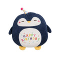 A plush penguin toy with a dark navy blue body, white belly, yellow feet and beak, and a small party hat. The belly is embroidered with 'HAPPY BIRTHDAY' in multicolored letters with sprinkle details, set against a plain light gray background.