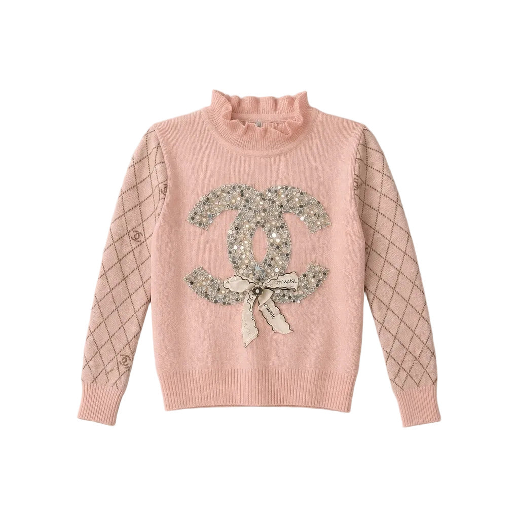 A pink knit sweater with a ruffled mock neck, featuring a large sequined Chanel logo with a bow on the chest and a subtle diamond pattern on the sleeves, laid flat on a light gray background.