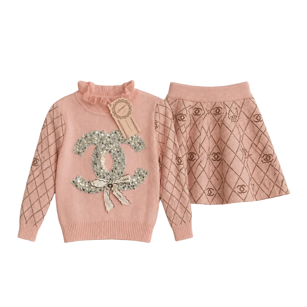 A pink knit children's sweater and matching skirt set laid flat on a light gray background. The sweater has a ruffled mock neck, silver rhinestone Chanel logo embellishment on the front with a bow detail, and dark geometric diamond patterns on the sleeves and hem.