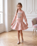 A young girl models a light pink two-piece knit set featuring a mock-neck long-sleeve top with a large silver Chanel logo on the chest and diamond-patterned sleeves, paired with a matching flared skirt. She is standing in a bright room with a herringbone wood floor and white paneled walls.