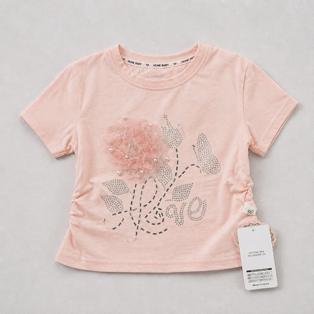 A light pink short-sleeve baby tee with a rhinestone floral design spelling 'love' and rhinestone leaves, featuring a ruffled flower applique on the chest. The brand 'JUNE BAPT' is visible on the inner neck label and hangtag.