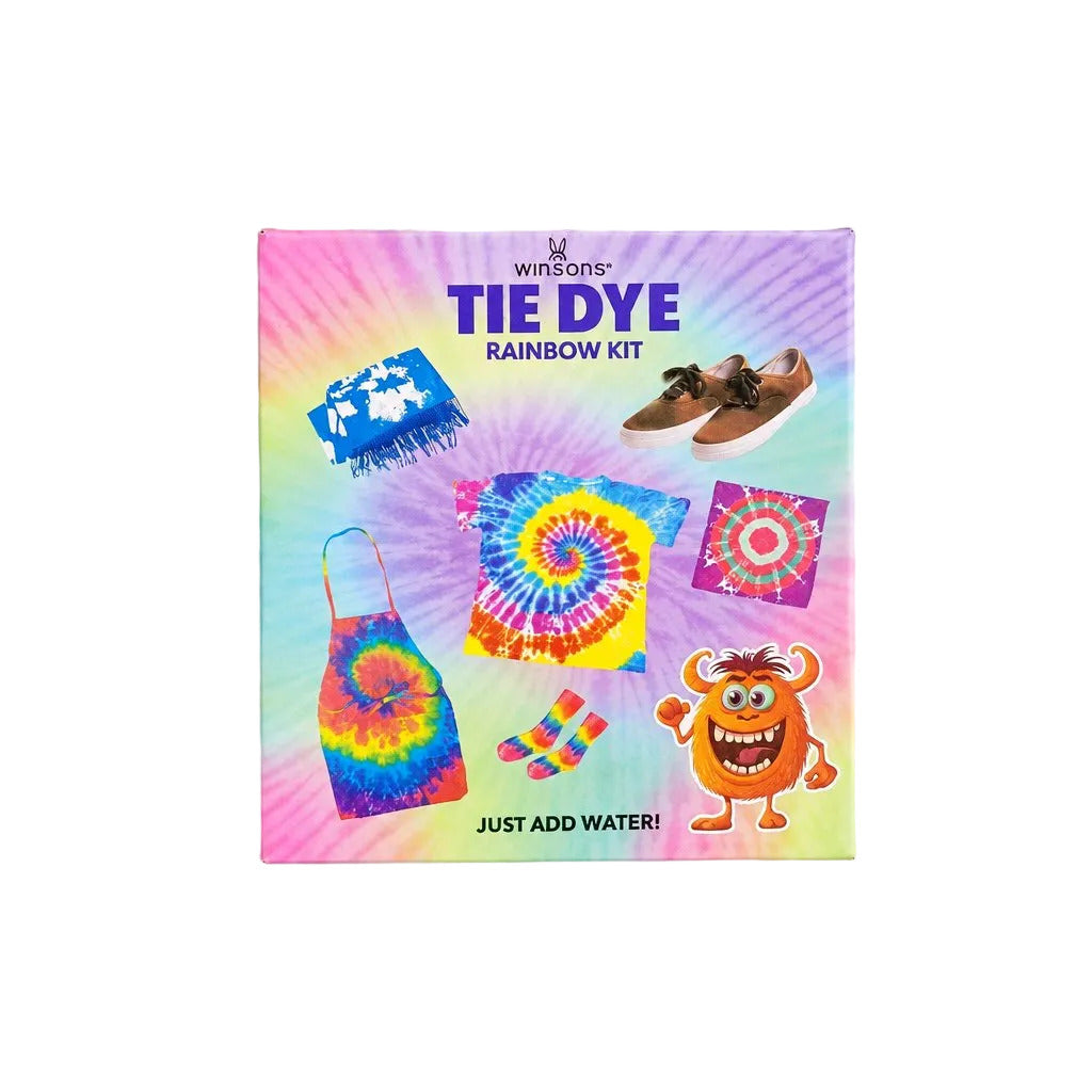 A square tie-dye rainbow kit box by We're a Tie-Dye Gang, featuring a colorful pastel rainbow design with examples of tie-dye patterns and accessories, photographed on a neutral light gray background.