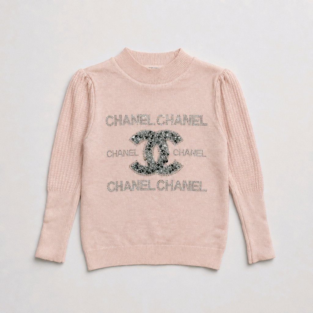 A light pink Chanel knit sweater with a textured waffle pattern, featuring a large Chanel logo and 'CHANELCHANEL' text in silver rhinestones on the front, laid flat against a light gray background.