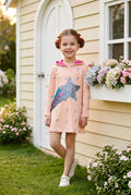 A young girl with pigtails stands in a garden wearing a light peach-pink hooded dress with a large blue and purple star design on the front. The dress has a pink hood lining and small gray bird prints.