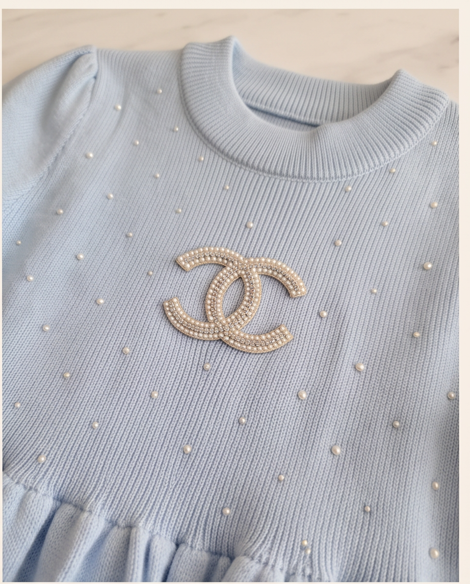 A light blue ribbed knit top with a cream Chanel logo brooch and small pearl-like dots, shown in a close-up product shot.