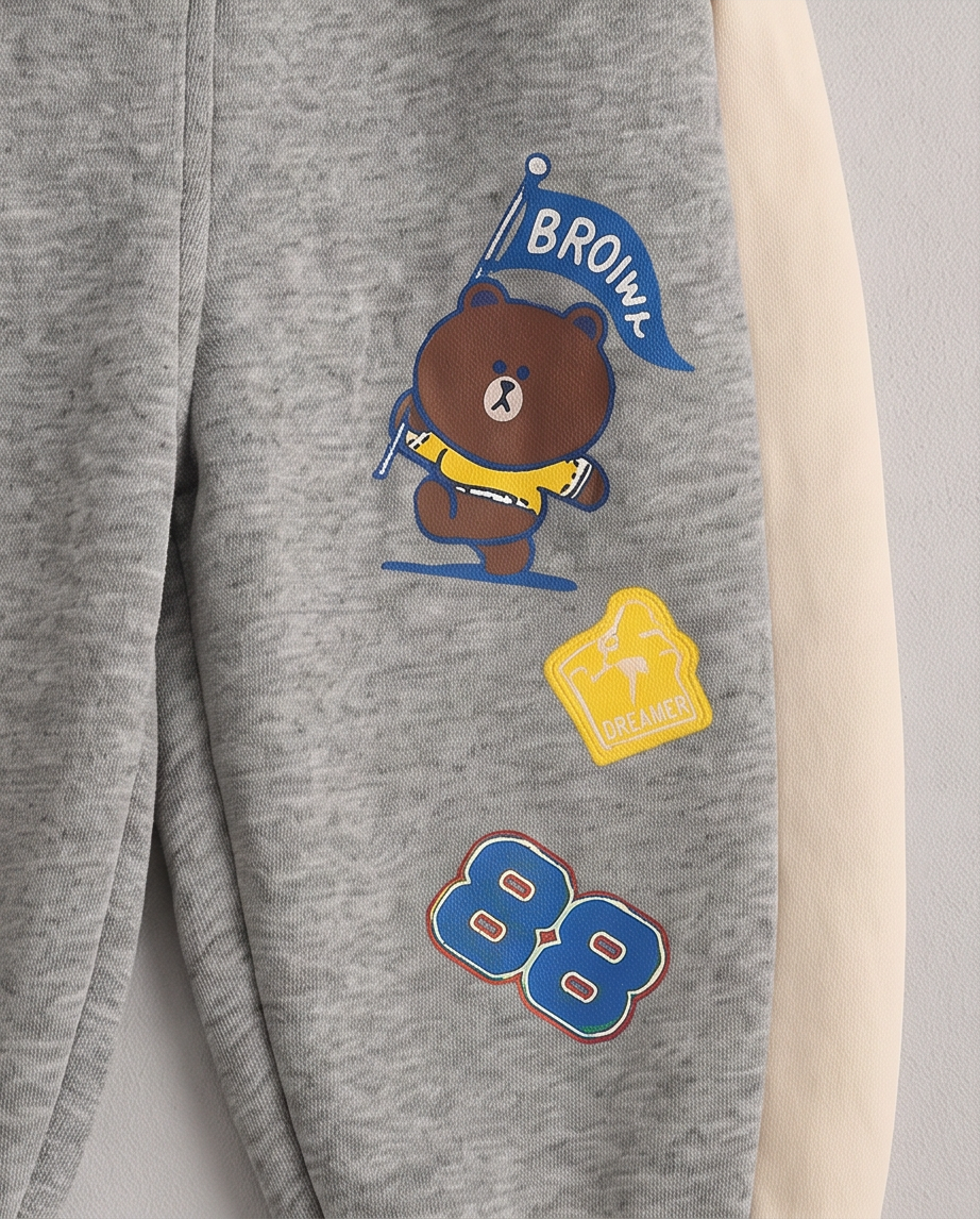 Close-up of heather gray sweatpants with a cream-colored side panel. The left leg features a graphic of the cartoon bear Brown holding a blue flag that says 'BROWN' in white, with a yellow 'DREAMER' patch below it and blue number 88 graphics near the bottom.
