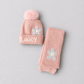 A pink knit baby hat with a fluffy pom-pom and the name 'Laucy' embroidered on the cuff, shown next to a matching pink knit bootie sock with a flower applique, on a light gray background.
