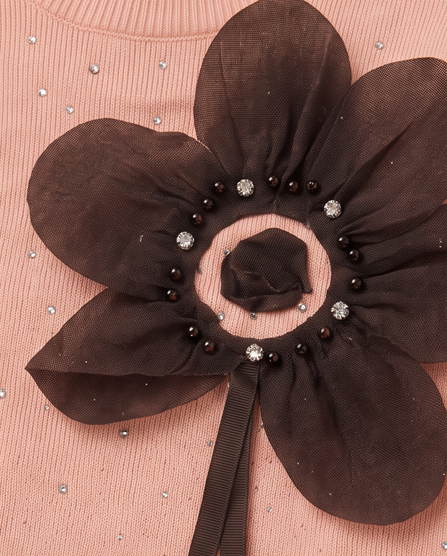 A close-up of a dark brown sheer organza flower applique embellished with black beads, crystal-like rhinestones, and a matching dark ribbon, attached to a pink ribbed knit garment.