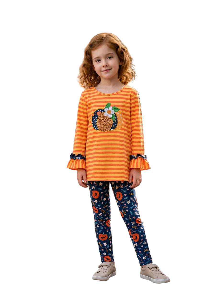 Young girl in an orange shirt with a pumpkin design and blue patterned leggings standing in an autumn park.