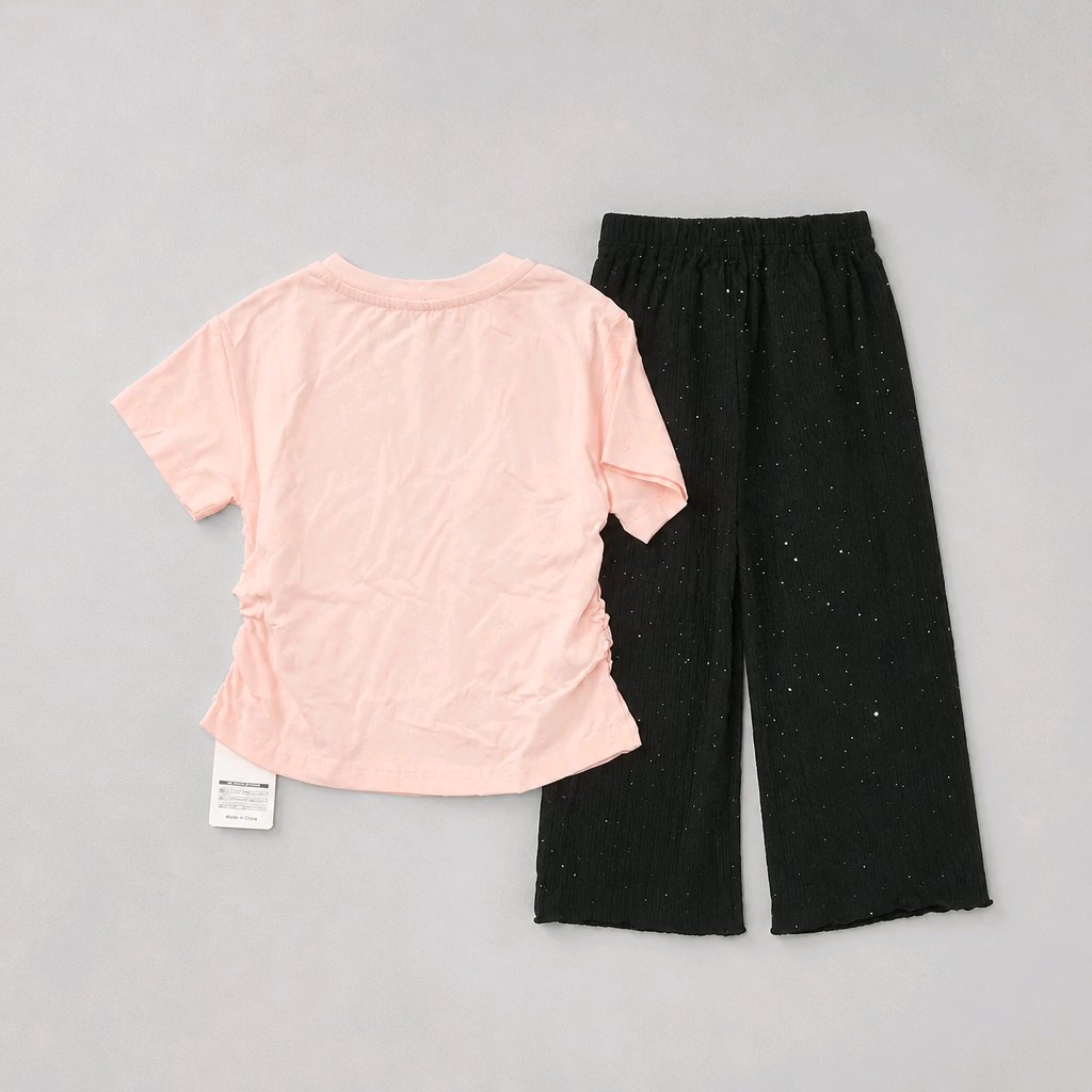 A two-piece children's outfit set featuring a light pink short-sleeve t-shirt with ruched side details and black wide-leg pants with subtle silver glitter speckles, laid flat on a neutral gray background.